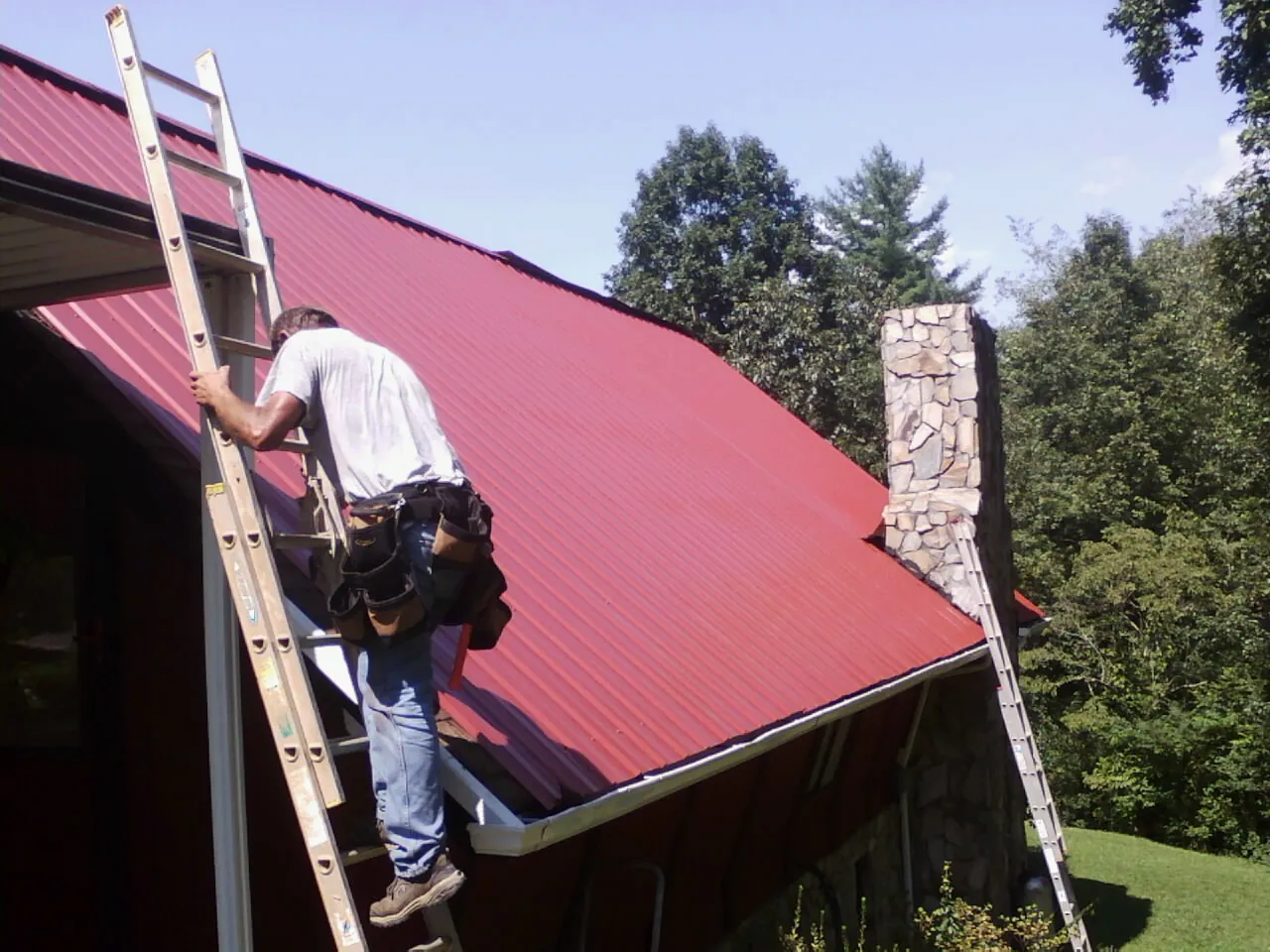 Roofer on a ladder beside a red metal roof and stone chimney — Soffit & Fascia Repair by Roofing Brothers in Eminence
